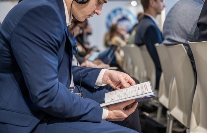 Participant in a suit reading the TRUSTECH 2025 conference program while wearing audio headphones in the conference room.