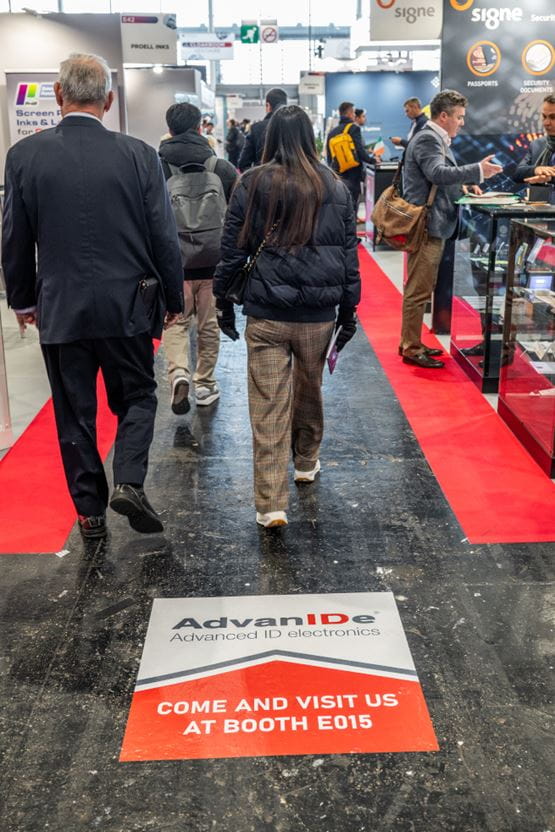 Allée du salon professionnel TRUSTECH avec un marquage au sol invitant les visiteurs à se rendre sur un stand spécifique.