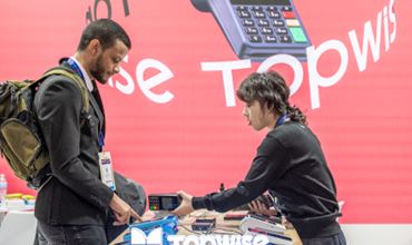 Démonstration d’un terminal de paiement sur un stand lors du salon TRUSTECH, entre une exposante et un visiteur.