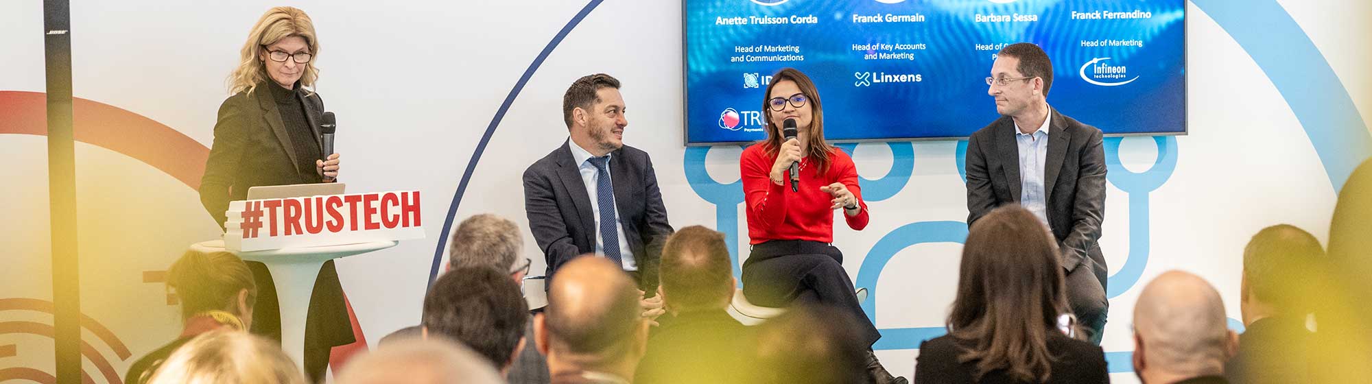 3 speakers sitting on the innovation stage speaking to an audience. A moderator stands on the left behind a lectern, microphone in hand, with a #trustech in the foreground.