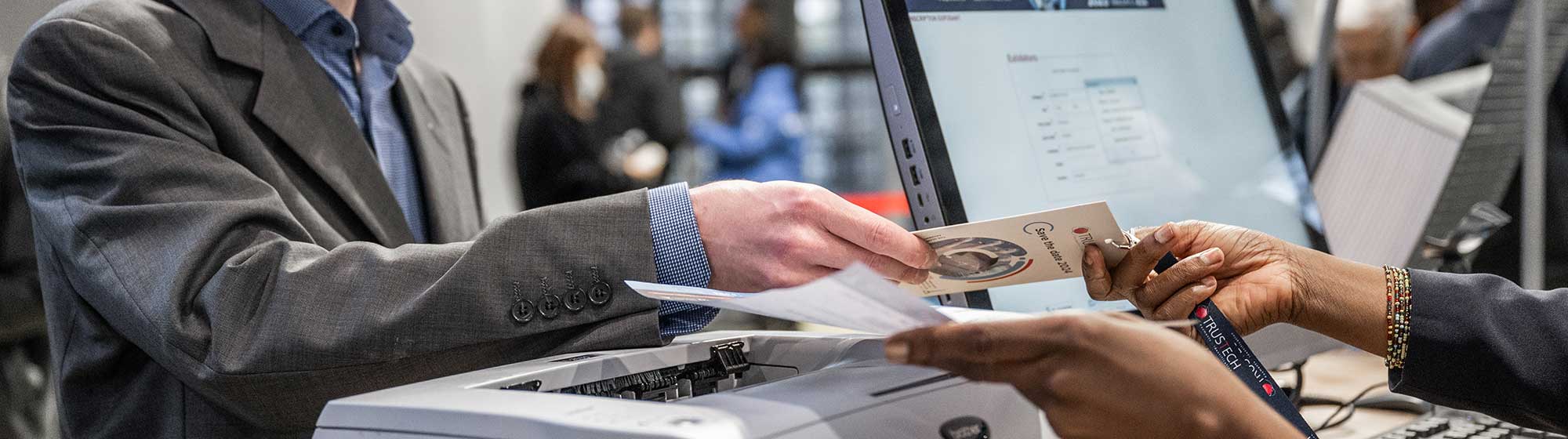 A visitor presents his pass to a trustech hostess. There is also a computer screen with the TRUSTECH registration page and a printer. 