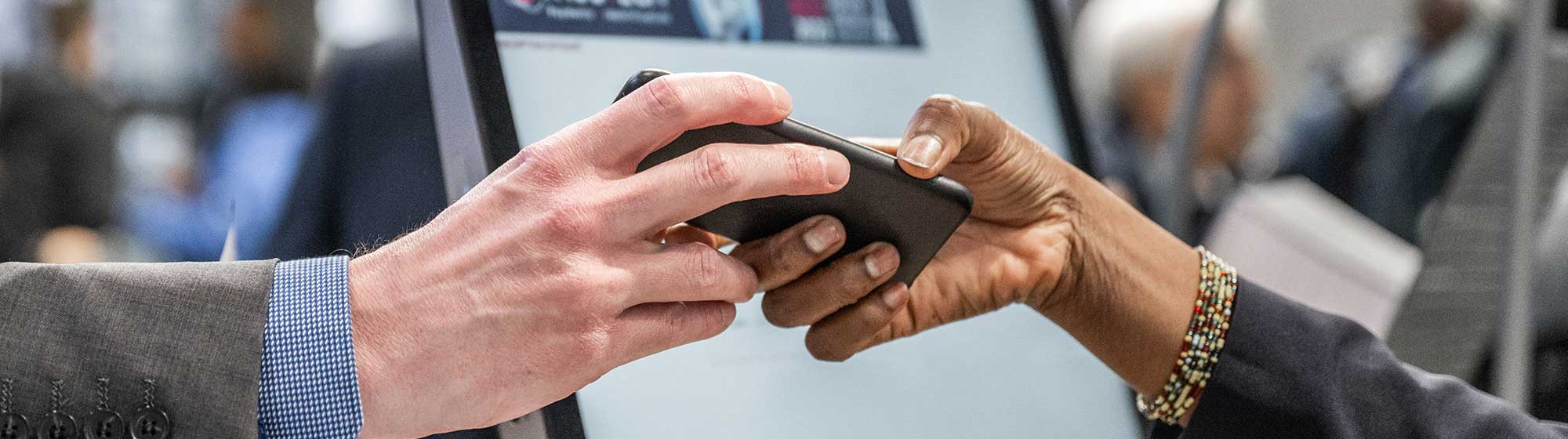 Two people are exchanging a smartphone in front of a computer screen. One person is handing the phone to another person at the TRUSTECH event. The computer screen displays a logo and some information, and there appear to be other people in the background.