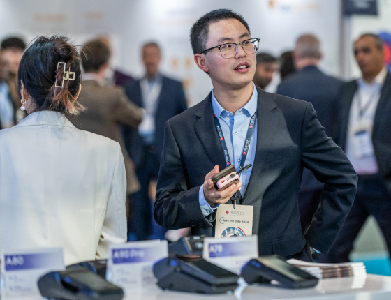 A businessman, with a Trustech badge around his neck, is looking at something off-screen, holding his phone in his hand. He is surrounded by a crowd of other visitors. Payment terminals are displayed in front of him.